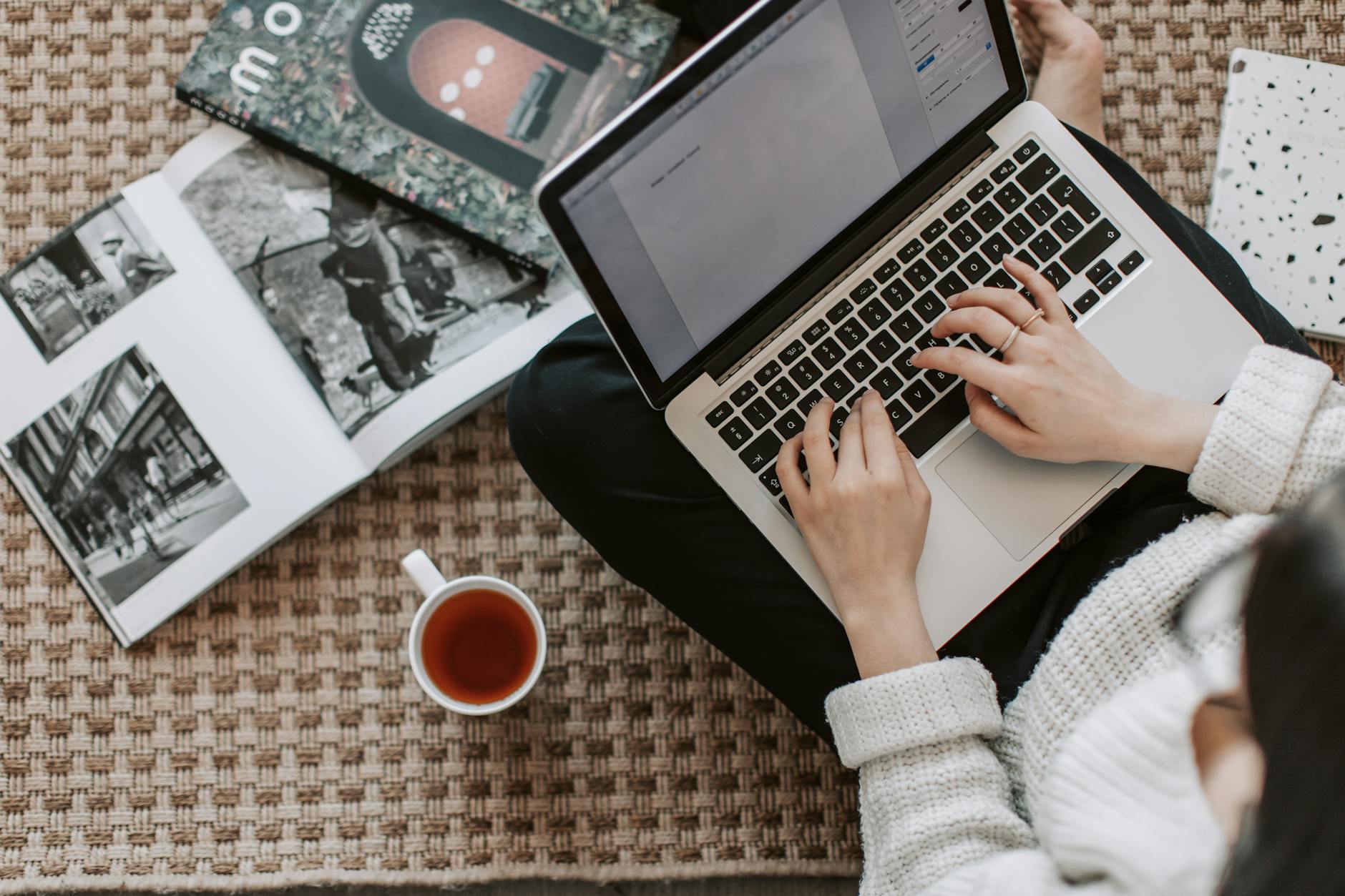 A person typing on a laptop while seated on a rug, surrounded by magazines and a cup of tea.